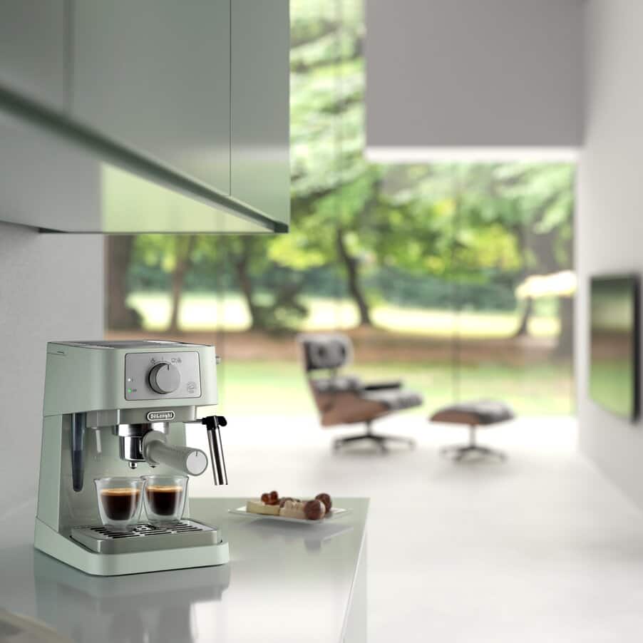 DeLonghi Stilosa EC260.GR espresso coffee machine in sage green and white dispensing two shots of espresso into glass cups on a white countertop, with modern interior and garden view in background