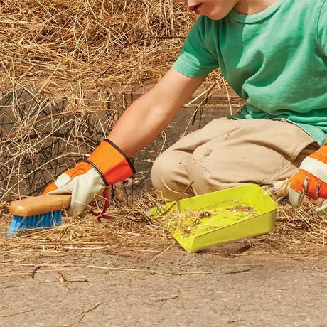 Child wearing green shirt and protective gloves using a blue brush and bright yellow dustpan to sweep straw and debris from ground outdoors