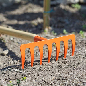 Bright orange children's garden rake with multiple curved tines positioned in loose soil, with a wooden handle visible in the background and garden beds out of focus