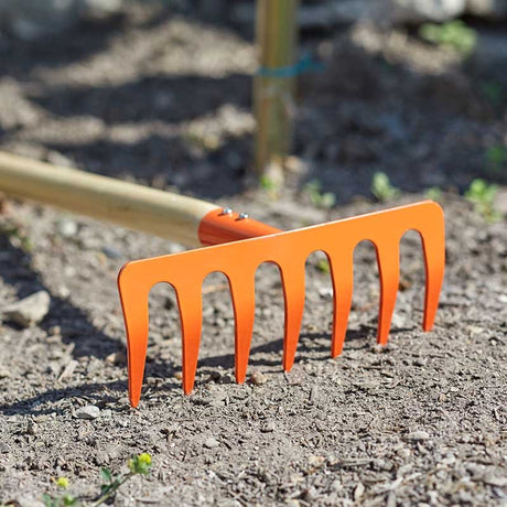 Bright orange children's garden rake with multiple curved tines positioned in loose soil, with a wooden handle visible in the background and garden beds out of focus