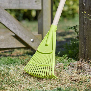 Bright lime green leaf rake with white handle resting on grass and soil near wooden garden fence, designed for children's gardening