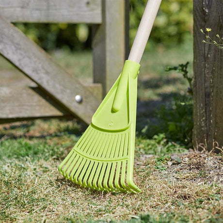 Bright lime green leaf rake with white handle resting on grass and soil near wooden garden fence, designed for children's gardening
