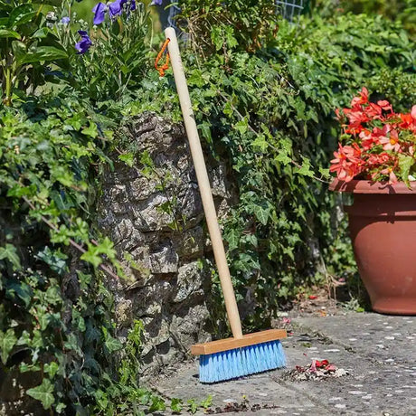 Child-sized garden sweeping brush with light blue bristles and wooden handle leaning against ivy-covered stone wall in garden setting with purple flowers, red potted flowers, and natural daylight