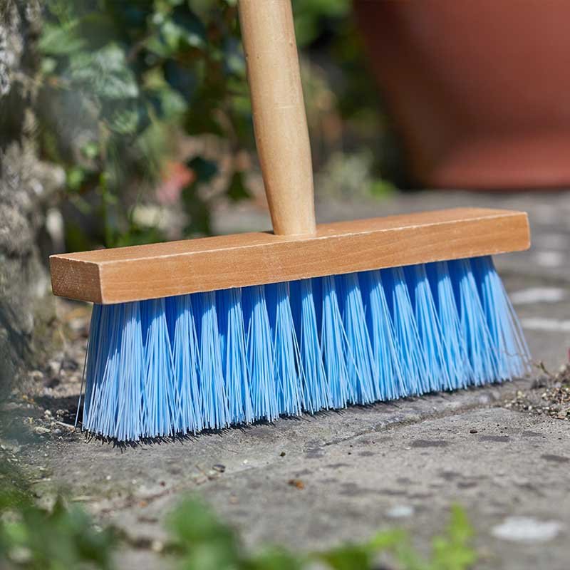 Child-sized sweeping brush with blue synthetic bristles and wooden handle resting on pavement, surrounded by garden foliage and terracotta pot