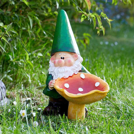 Garden gnome ornament with green pointed hat and white beard holding a red and white spotted mushroom, displayed on green grass with foliage background