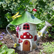 Whimsical mushroom-shaped garden watering can ornament with red and white spotted roof, metal construction, decorative windows and door, surrounded by green foliage and yellow flowers in a garden setting