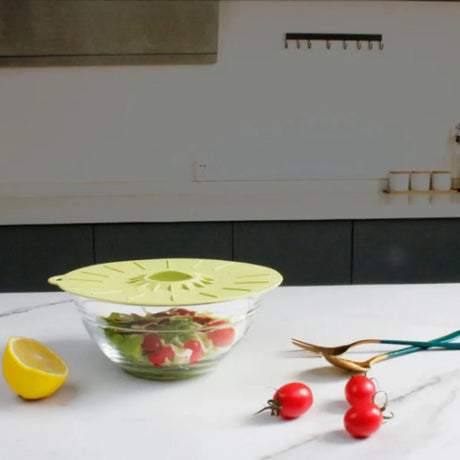 Yellow silicone suction lid covering a glass bowl of salad in a modern kitchen, with fresh tomatoes, lemon half, and kitchen shears on white countertop