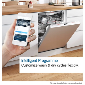 White hand holding smartphone displaying Bosch dishwasher app with wash cycle controls, positioned next to integrated Bosch Series 6 dishwasher with beige door panel in modern white kitchen with wooden countertop, demonstrating intelligent programme customization feature