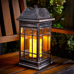 Smart Solar Seville solar lantern with dark metal frame and amber glass panels emitting warm yellow light, positioned on wooden garden furniture at night with foliage visible in background
