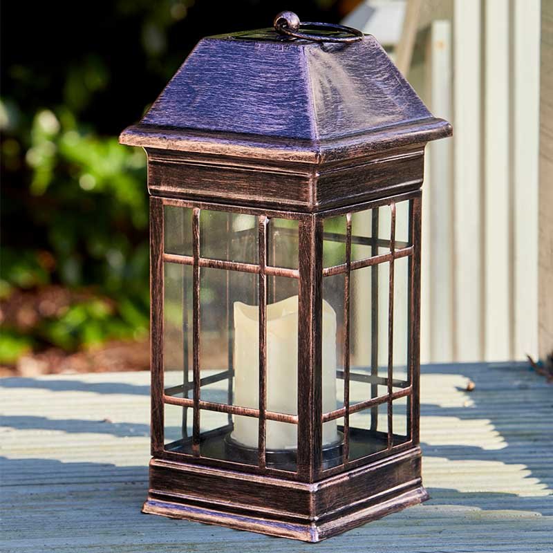 Solar-powered Seville lantern with aged bronze finish, grid-patterned glass panels, and pyramid roof displayed on wooden deck with garden background
