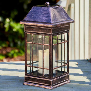 Solar-powered Seville lantern with aged bronze finish, grid-patterned glass panels, and pyramid roof displayed on wooden deck with garden background