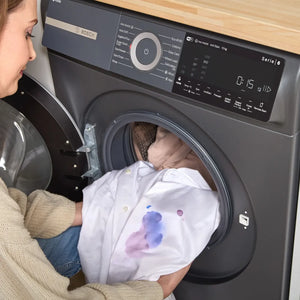 Person loading white shirt with blue and purple stains into open drum of Bosch Series 6 grey front-load washing machine with digital control panel displaying time and settings