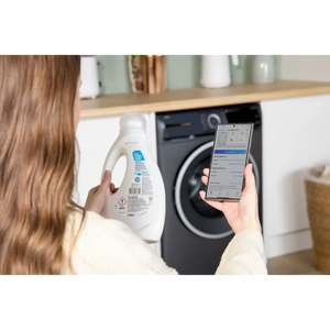 Woman holding smartphone displaying washing machine app controls next to grey Bosch Series 6 front-load washing machine in modern white and wood kitchen with detergent bottle
