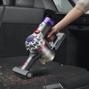 Dyson V8 Absolute cordless stick vacuum with purple and red accents being used to clean a car interior, showing the handheld attachment with motorized brush head and dust cup