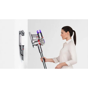 Woman in white long-sleeve shirt holding silver and purple Dyson V8 Advanced cordless stick vacuum cleaner with wall-mounted charging dock shown on left side
