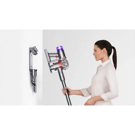 Woman in white long-sleeve shirt holding silver and purple Dyson V8 Advanced cordless stick vacuum cleaner with wall-mounted charging dock shown on left side