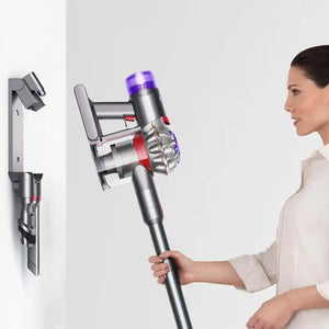 Woman in white shirt holding silver Dyson V8 Absolute cordless stick vacuum with purple LED display and red accents, with wall-mounted charging dock visible on left side
