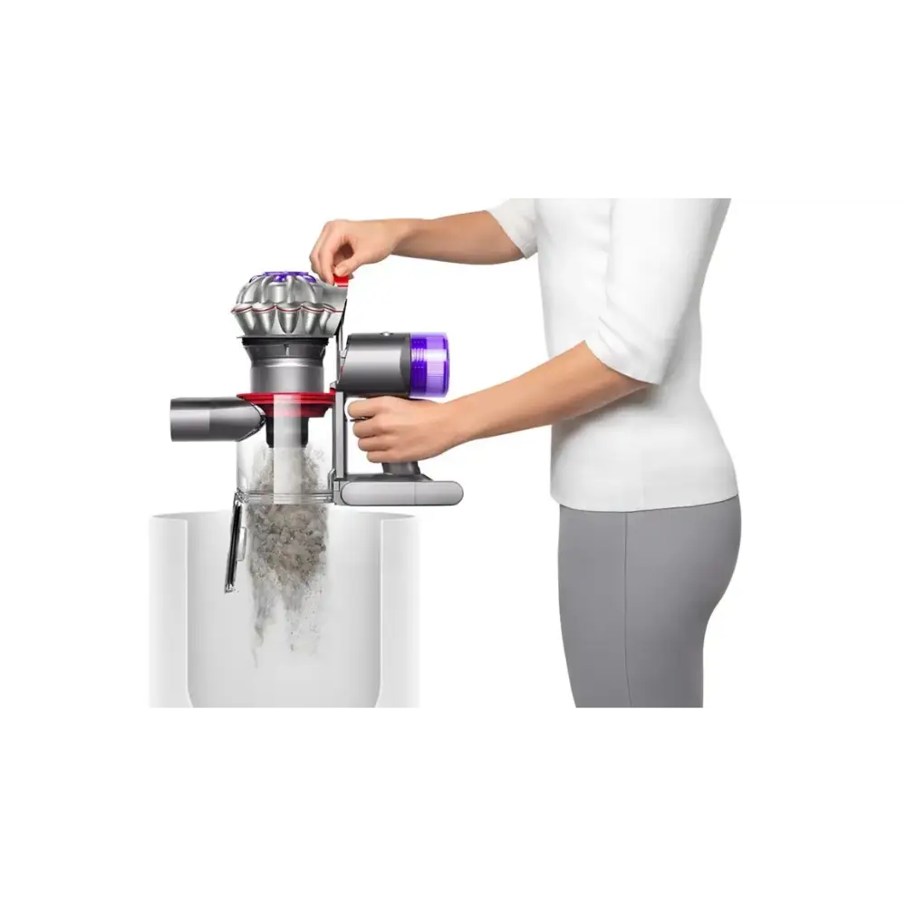 Woman in white top demonstrating Dyson V8 Advanced cordless stick vacuum with purple filter head being removed over white collection bin, showing dust emptying mechanism