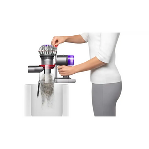 Woman in white top demonstrating Dyson V8 Advanced cordless stick vacuum with purple filter head being removed over white collection bin, showing dust emptying mechanism
