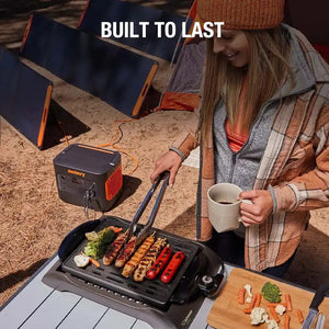 Woman in brown knit beanie and plaid jacket grilling food on portable electric griddle powered by Jackery portable power station and solar panels, camping setup with mug and prepared vegetables on wooden cutting board
