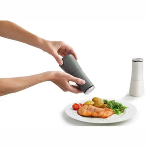 Hands sprinkling black pepper from gray Joseph Joseph Milltop grinder onto plated salmon dinner with potatoes, tomatoes and greens, white salt shaker visible beside plate