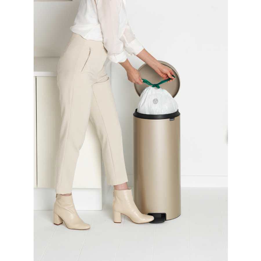 Woman in cream trousers and white shirt disposing of waste into a champagne-colored stainless steel Brabantia pedal bin with black foot pedal and rim, showing the bin's sleek cylindrical design and ease of use