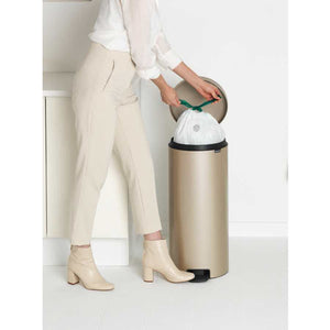Woman in cream trousers and white shirt disposing of waste into a champagne-colored stainless steel Brabantia pedal bin with black foot pedal and rim, showing the bin's sleek cylindrical design and ease of use
