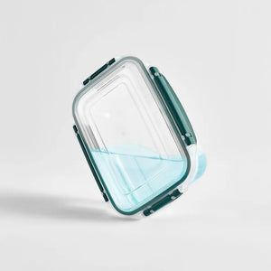 Clear glass rectangular storage container with teal silicone handles and locking clips, partially filled with light blue liquid, tilted at an angle against a white background