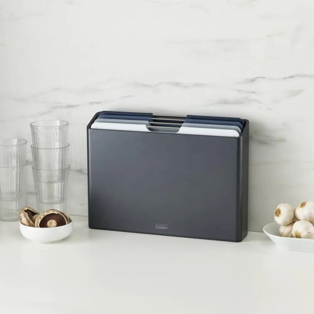Joseph Joseph Folio 4 Piece Chopping Board Set in graphite displayed on a marble countertop with stacked glass cups and bowls of mushrooms and onions