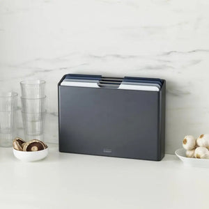 Joseph Joseph Folio 4 Piece Chopping Board Set in graphite displayed on a marble countertop with stacked glass cups and bowls of mushrooms and onions