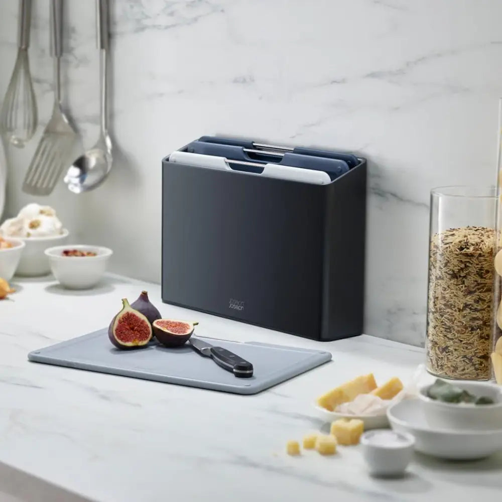 Joseph Joseph Folio 4 Piece Chopping Board Set in graphite displayed on a marble kitchen countertop with fresh figs, cereals, and cooking utensils