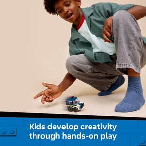 Young child in green cardigan and blue socks crouching down to play with a small blue and white LEGO police truck toy on a beige surface, demonstrating hands-on creative play