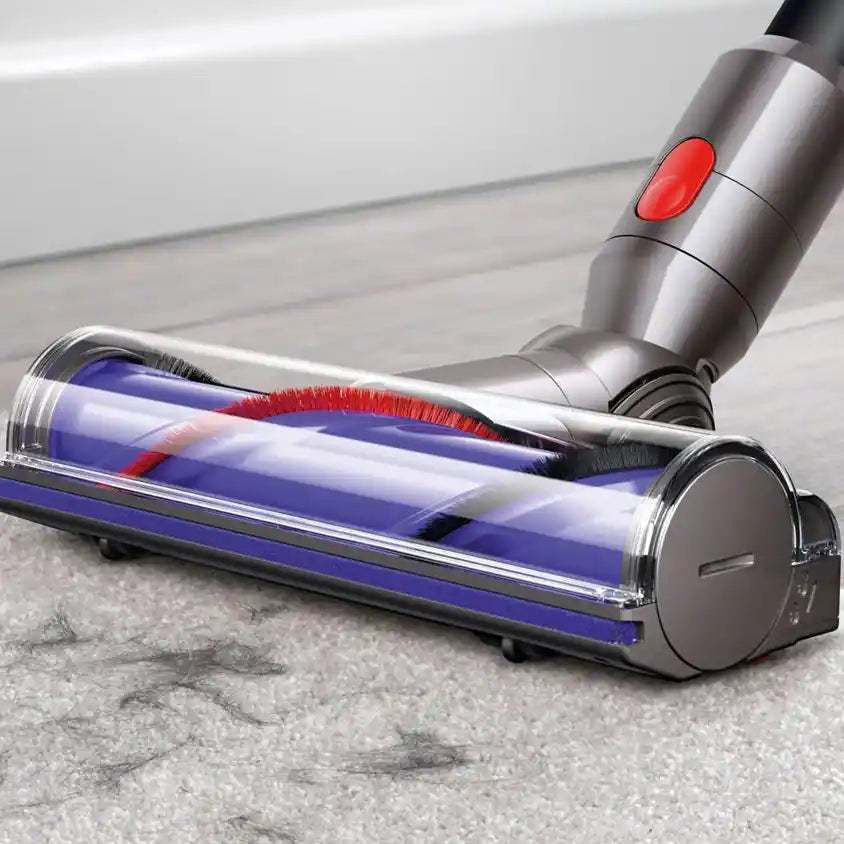 Dyson V8 Absolute cordless stick vacuum with silver metallic body and red power button cleaning gray carpet, showing purple dust bin and motorized brush head in action