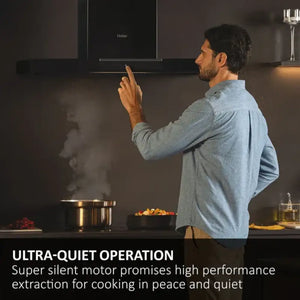 Man in light blue shirt using touch control on Haier black wall-mounted chimney cooker hood in modern kitchen with stainless steel pot on induction hob, demonstrating ultra-quiet operation feature