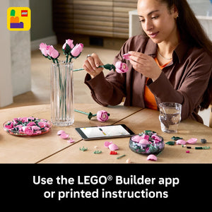 Woman building LEGO pink roses at wooden table with LEGO Builder app on smartphone, displaying assembled flower stems in glass vase and completed rose arrangements in decorative bowls