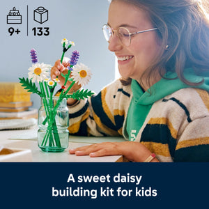 Young girl wearing glasses and a green and cream striped sweater smiling while admiring a LEGO Flowers daisy building set displayed in a clear glass vase, featuring white daisies with yellow centers and purple flowers, representing the 133-piece construction kit recommended for ages 9 and up