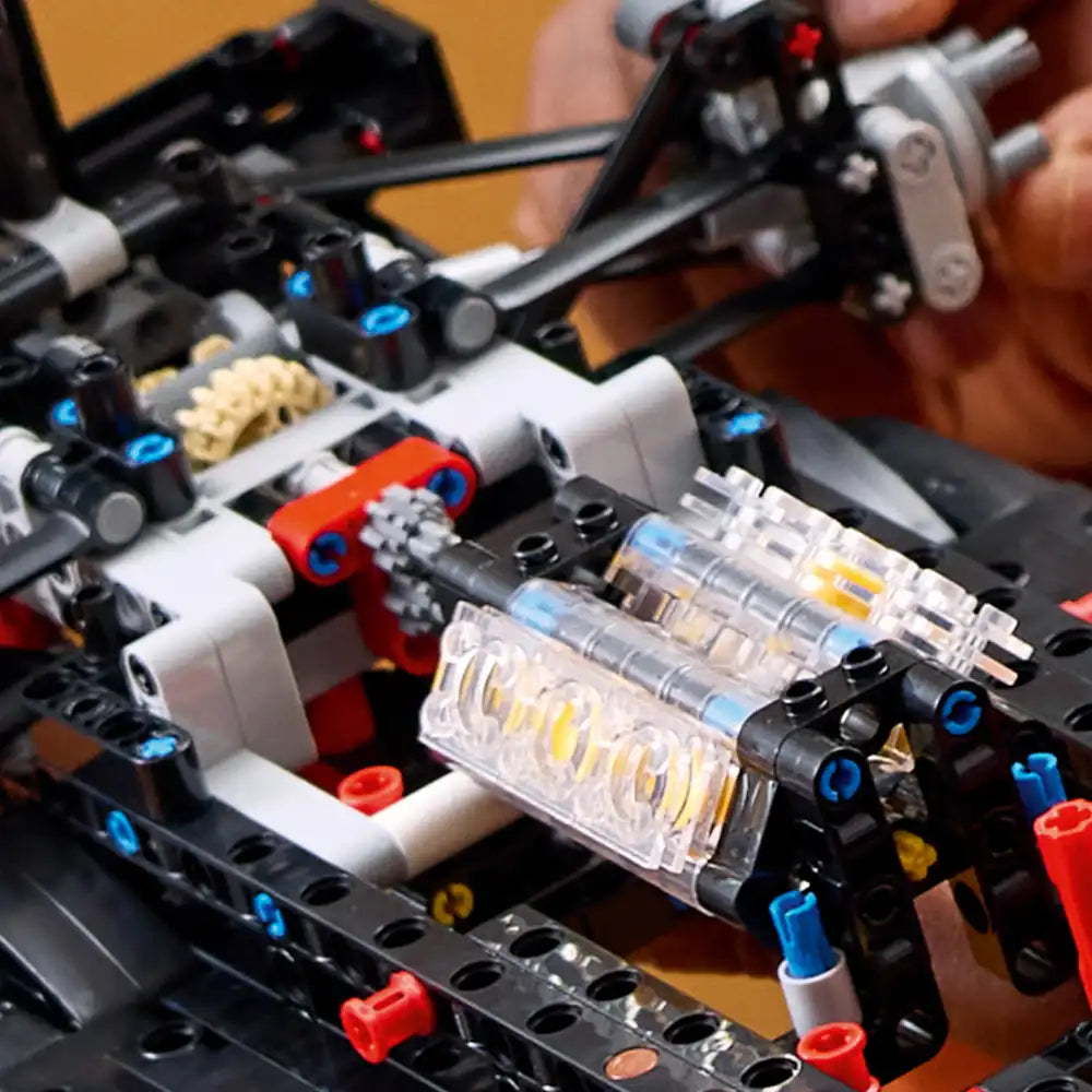 Close-up of LEGO Technic Mercedes-AMG F1 W14 E Performance model showing detailed engine assembly with transparent cylindrical components, blue and red mechanical parts, white connectors, and black chassis frame during construction