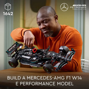 Man in orange sweater assembling black Mercedes-AMG F1 W14 E Performance Lego Technic model with PETRONAS branding at wooden table