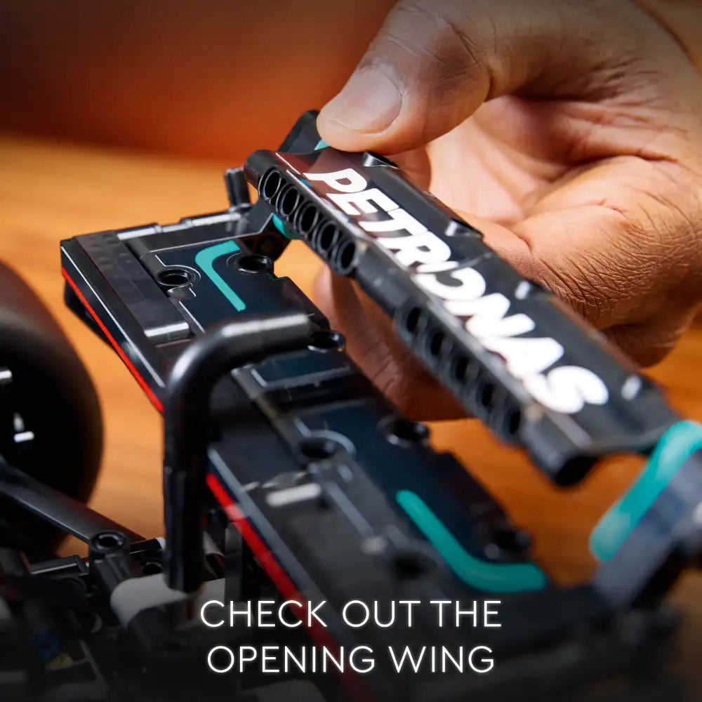 Hand holding a black LEGO Technic Mercedes-AMG F1 W14 E Performance model showing the detailed opening wing mechanism with turquoise and red accents on an orange background