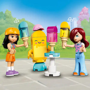 LEGO Friends ice cream and balloon stand set 42692 featuring two minifigure characters with colorful ice cream cone pieces in yellow, pink, and blue, a smiling yellow ice cream machine, and carnival-style balloon stand accessories on a gray paved surface with green landscaping background