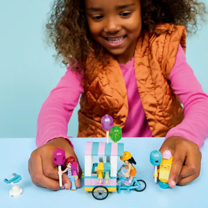 Child playing with LEGO Friends Ice Cream and Balloon Stand set 42692, featuring a colorful ice cream cart with pink and white striped awning, yellow bicycle, minifigure characters, and purple and blue balloons against a light blue background