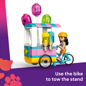 LEGO Friends Ice Cream and Balloon Stand set 42692 featuring a colorful turquoise pink and yellow ice cream cart with dispensers and three pink balloons on top, alongside a female minifigure in orange outfit and yellow helmet sitting on a turquoise bicycle attached to the stand