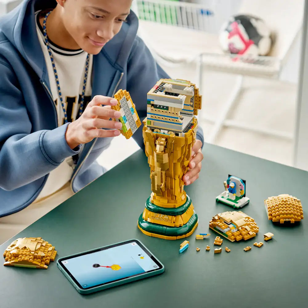 Child in blue jacket assembling gold and green LEGO FIFA World Cup Trophy set on table with instruction manual and loose brick pieces displayed
