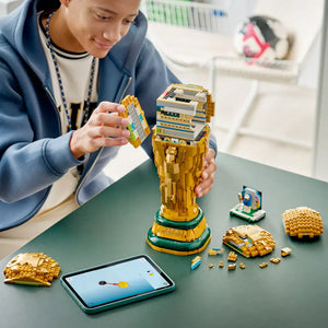 Child in blue jacket assembling gold and green LEGO FIFA World Cup Trophy set on table with instruction manual and loose brick pieces displayed