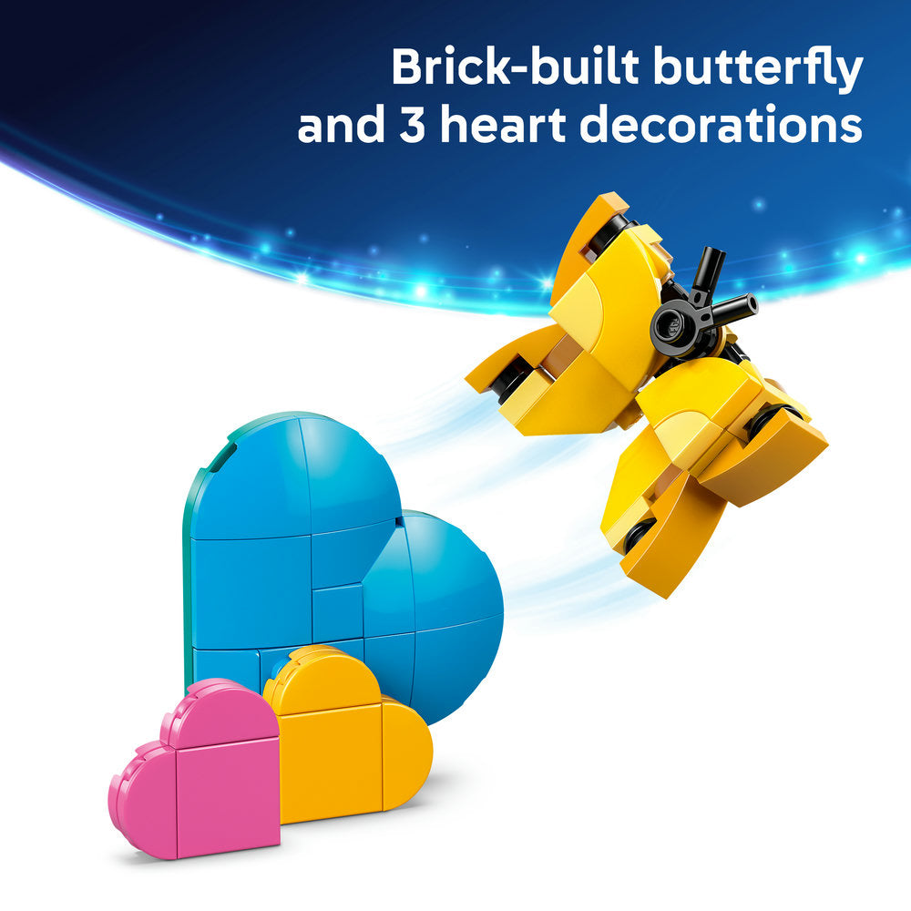LEGO brick-built yellow butterfly figure with articulated wings hovering above three heart decorations in blue, yellow, and pink, displayed against a blue gradient background with light effects