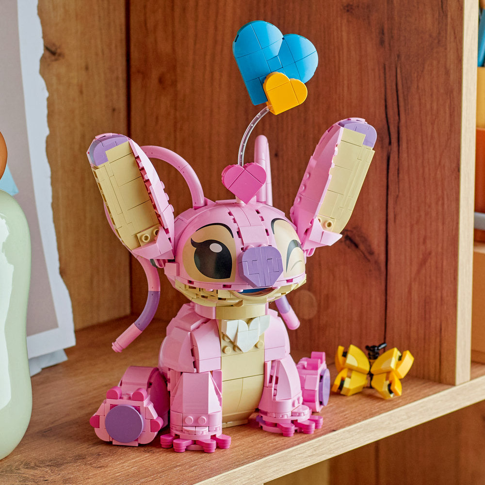 LEGO buildable pink Angel character figure with cream-colored chest and face, purple accents, articulated arms raised upward, holding blue and yellow heart-shaped balloons above head, displayed on wooden shelf with wooden backdrop