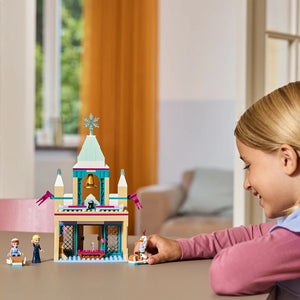 Young girl playing with LEGO Arendelle Castle set featuring blue and cream colored castle structure with snowflake topper, purple flags, and minifigures including Elsa and Anna characters in a bright living room setting