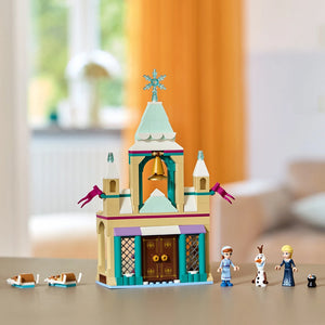 LEGO Arendelle Castle set 43265 featuring a turquoise and cream colored chapel with a cross on top, gold bell, purple flags, brown doors, and minifigures including Elsa, Anna, Olaf and a cat, displayed with loose brown brick elements on a surface with blurred background