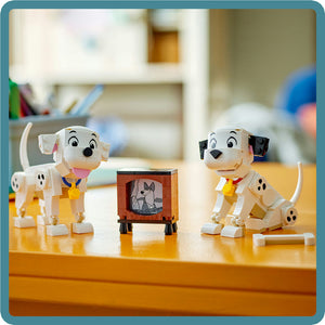 LEGO 101 Dalmatians Lucky and Penny white puppy figurines with yellow collars positioned on wooden surface beside small brown television set displaying dalmatian character, playroom background with shelving and toys