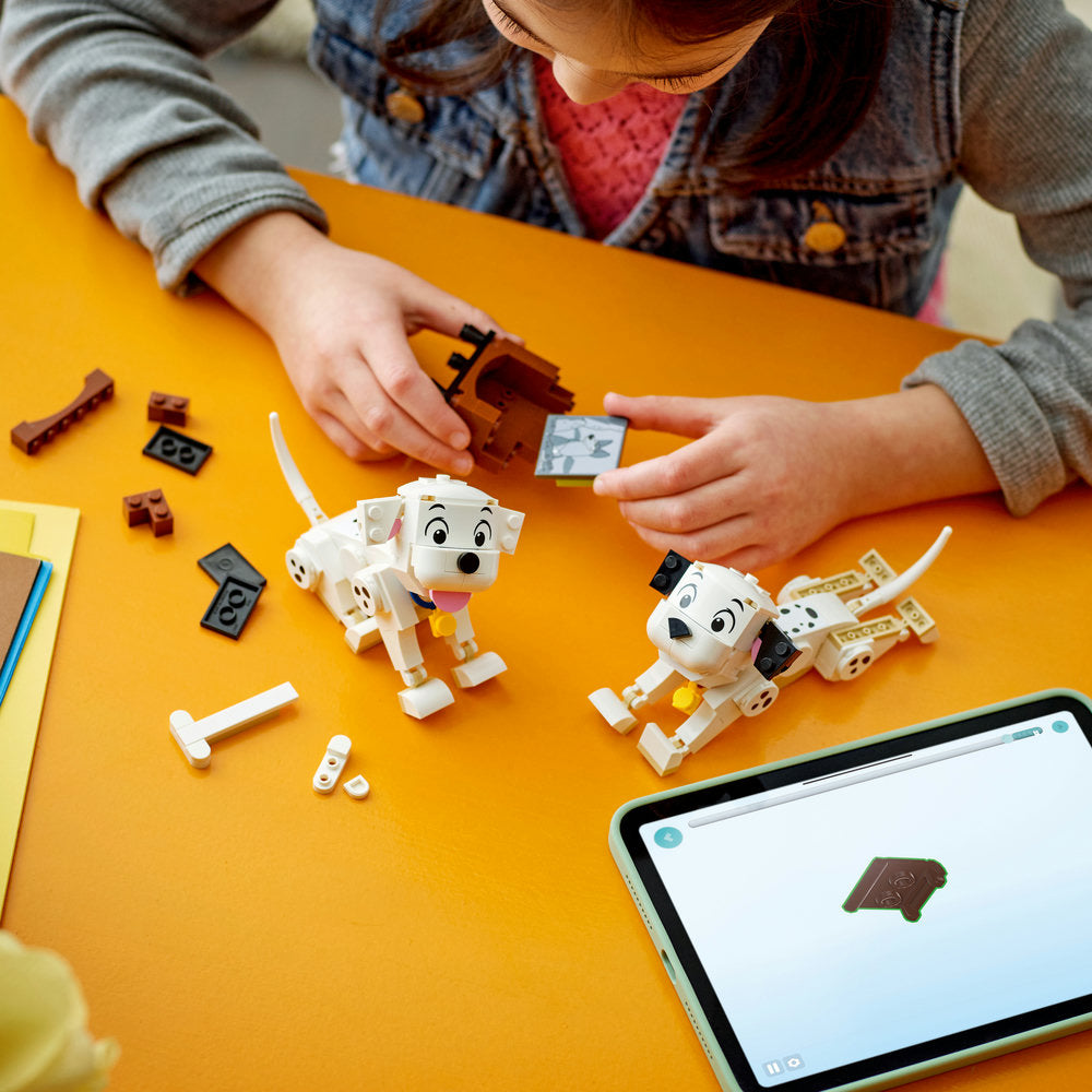 Child assembling LEGO 101 Dalmatians Lucky and Penny puppy figures on bright yellow surface with scattered LEGO bricks, instruction manual and tablet displaying building guide